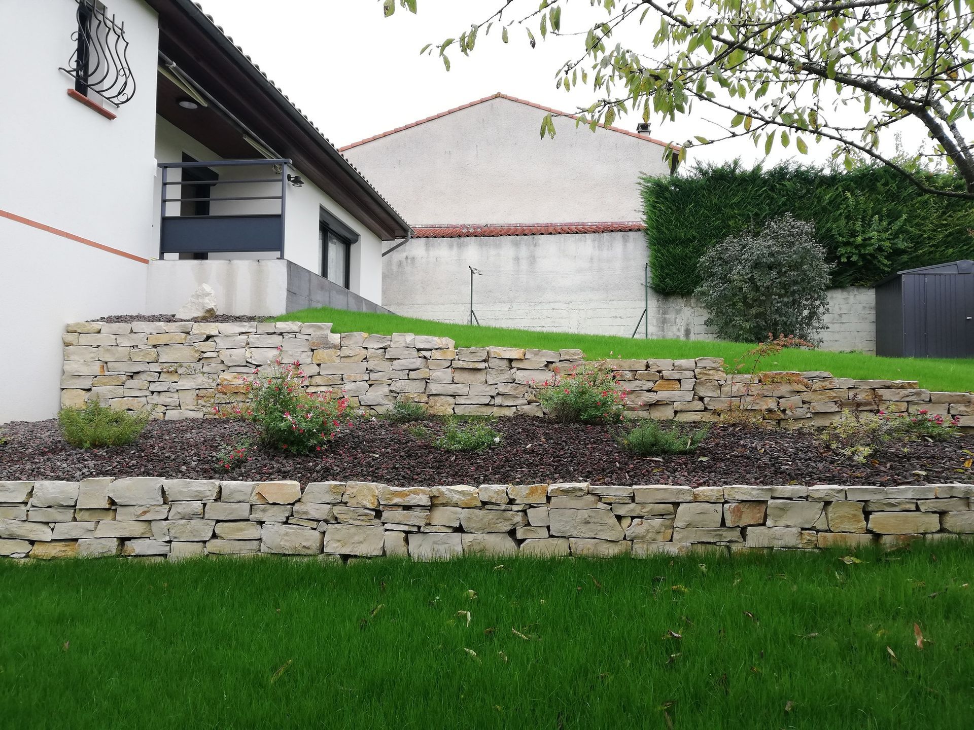Des murets de soutènement en pierre à plusieurs niveaux créent des parterres de jardin avec du paillis et des arbustes dans une cour arrière gazonnée jouxtant une maison.