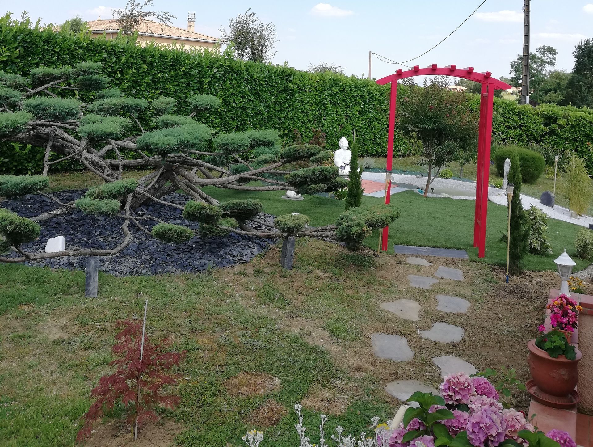 Un jardin avec un grand arbre de style bonsaï, une arche en bois rouge enjambant un chemin de pierre et une statue blanche sur une pelouse verte.