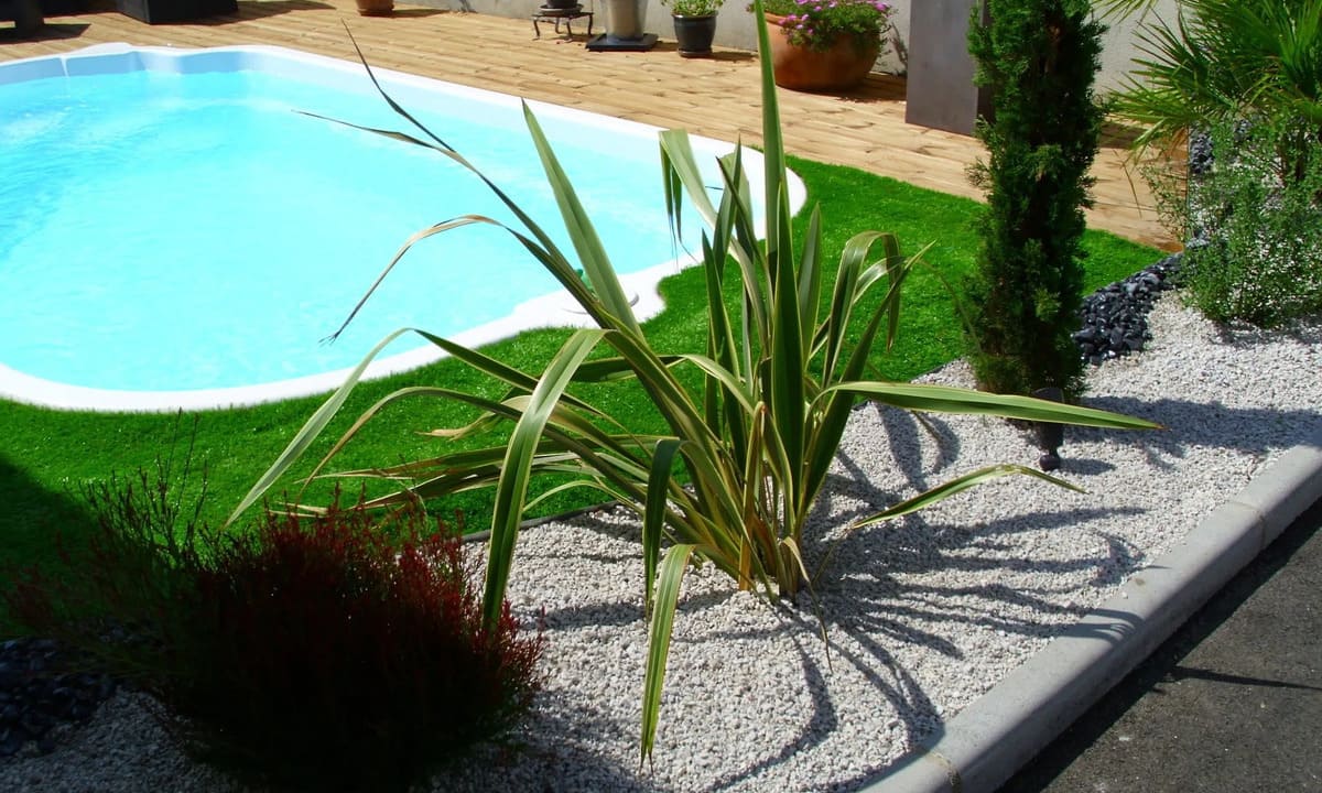 Une piscine bleu clair intégrée à un jardin paysager avec pelouse verte, gravier clair et plantes variées.