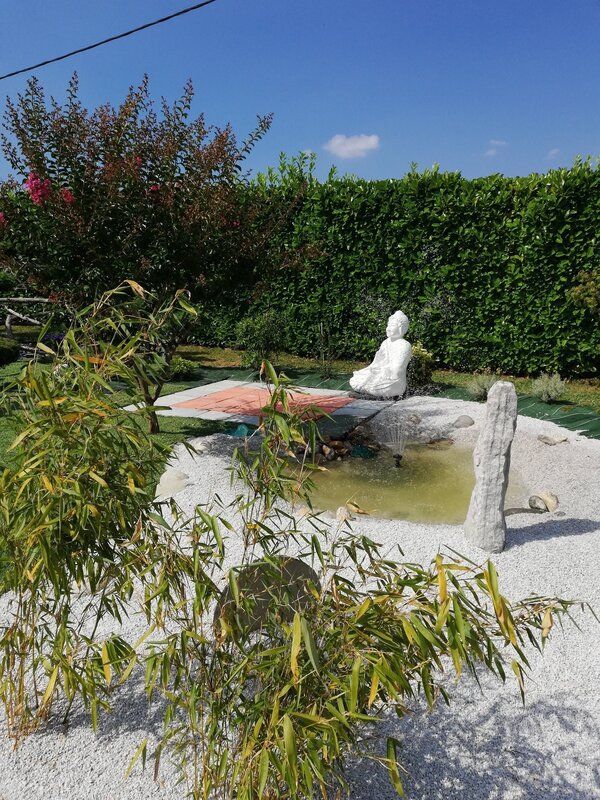 Une statue de Bouddha blanche se dresse près d'un petit étang dans un jardin zen, avec des bambous, du gravier blanc et une haie verte en arrière-plan.