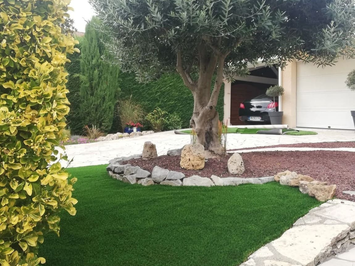 Un olivier se dresse dans un jardin recouvert de gazon synthétique, de pierres décoratives et de paillis rouge, près de l'allée d'une maison.