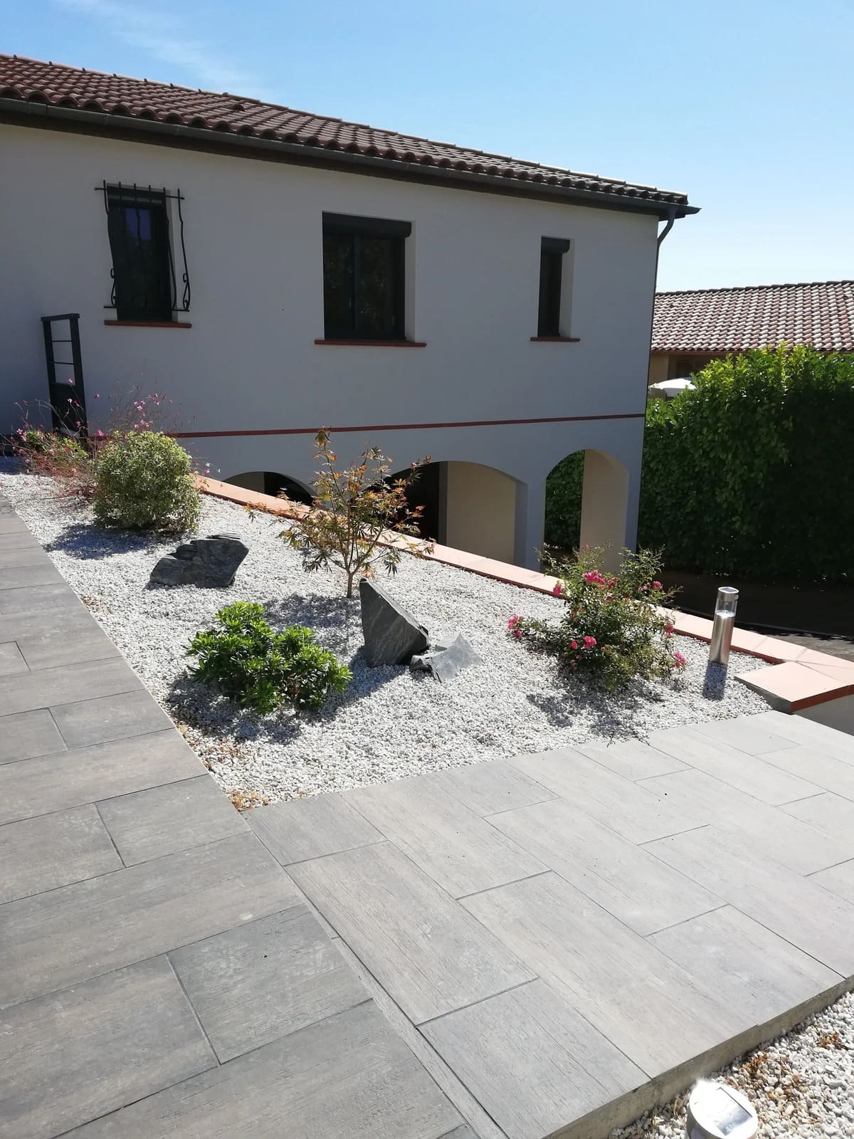 Une maison de deux étages aux tons clairs, avec une terrasse en pierre grise et un parterre de jardin rempli de gravier clair et de petits arbustes.