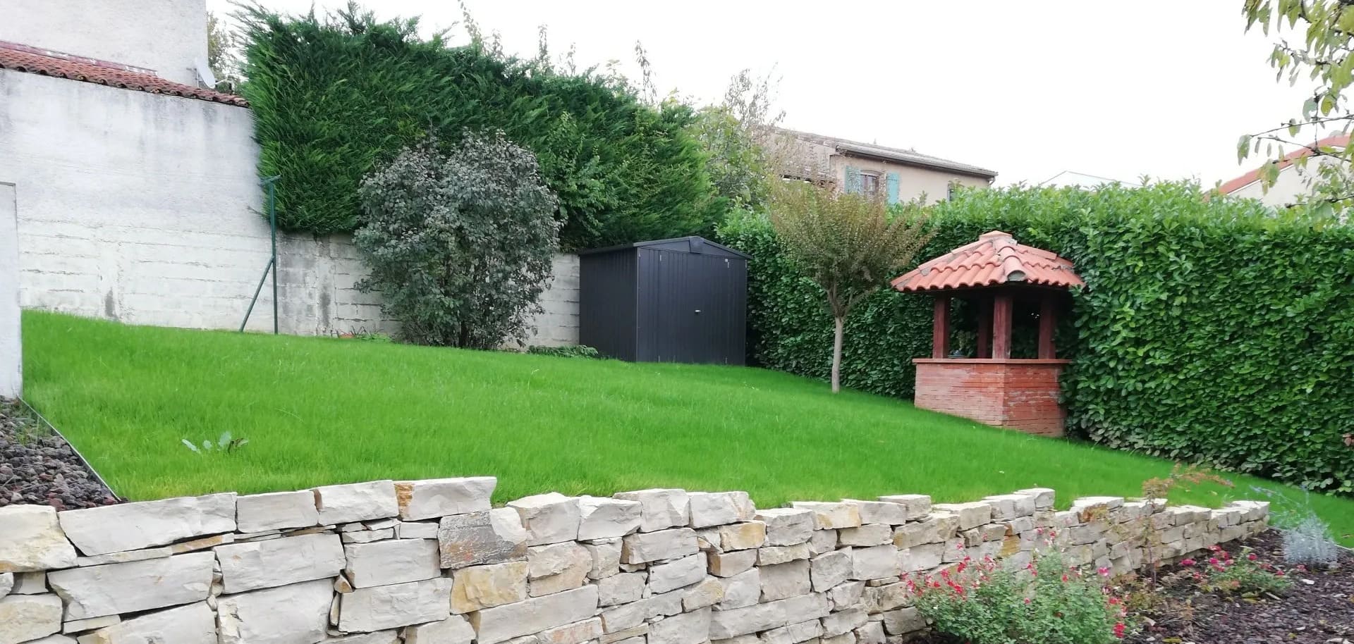 Dans un jardin, une pelouse en pente est agrémentée d'un muret de soutènement en pierre, d'un petit abri noir et d'un puits décoratif en briques.