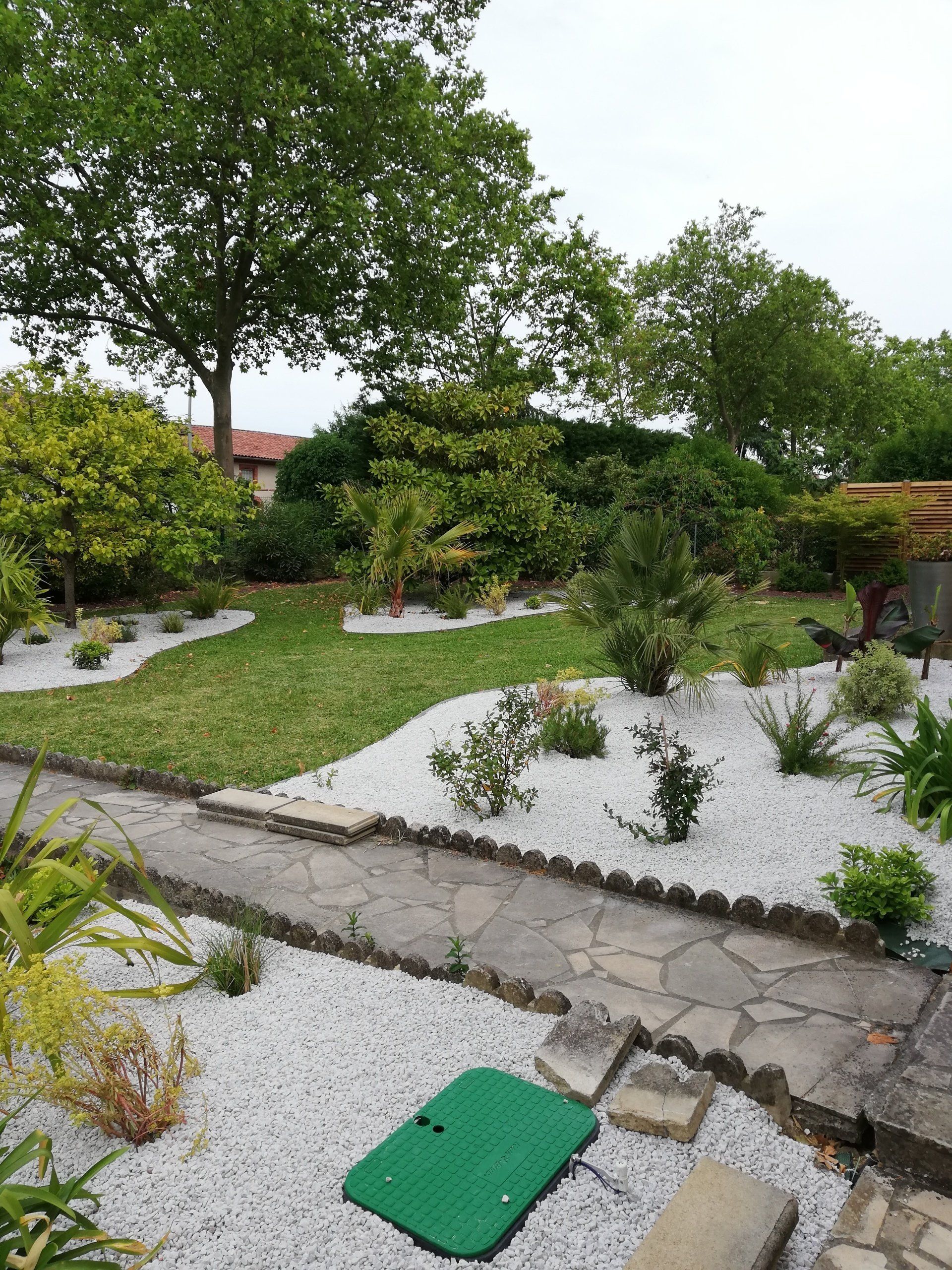 Un jardin luxuriant est agrémenté d'un chemin de pierre serpentant entre des zones de gravier blanc, d'herbe verte, d'arbustes et de grands arbres.
