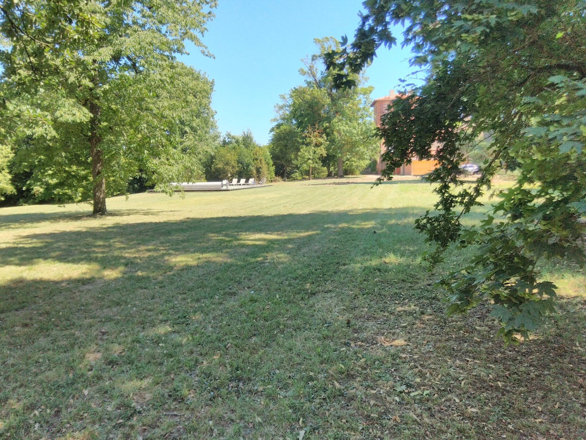Un champ herbeux bordé d'arbres matures par une journée ensoleillée, avec au loin une maison et du mobilier de jardin.