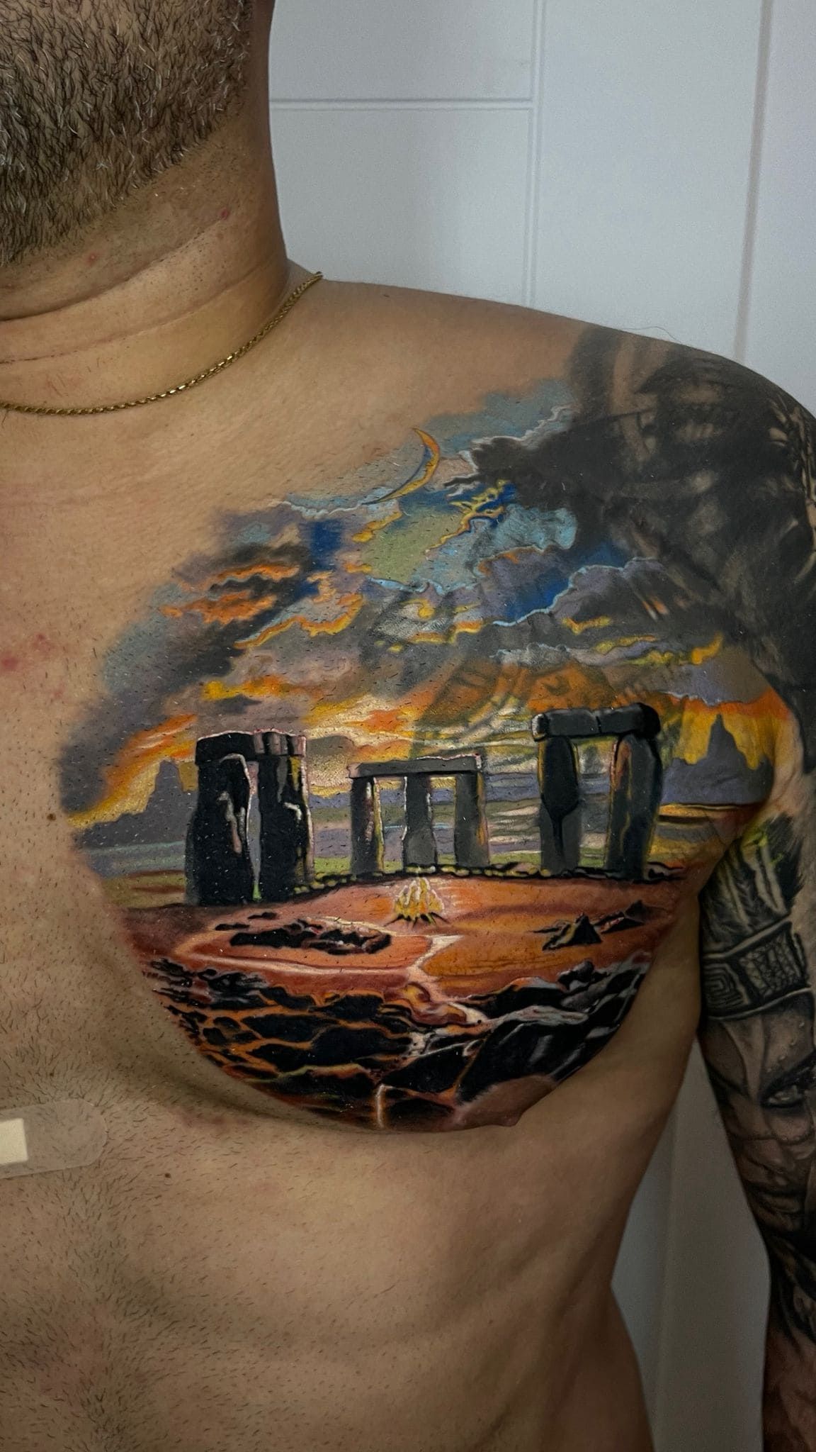 Tatuaje en el hombro de un hombre que representa un paisaje al atardecer con pilares de piedra oscura y nubes naranjas.