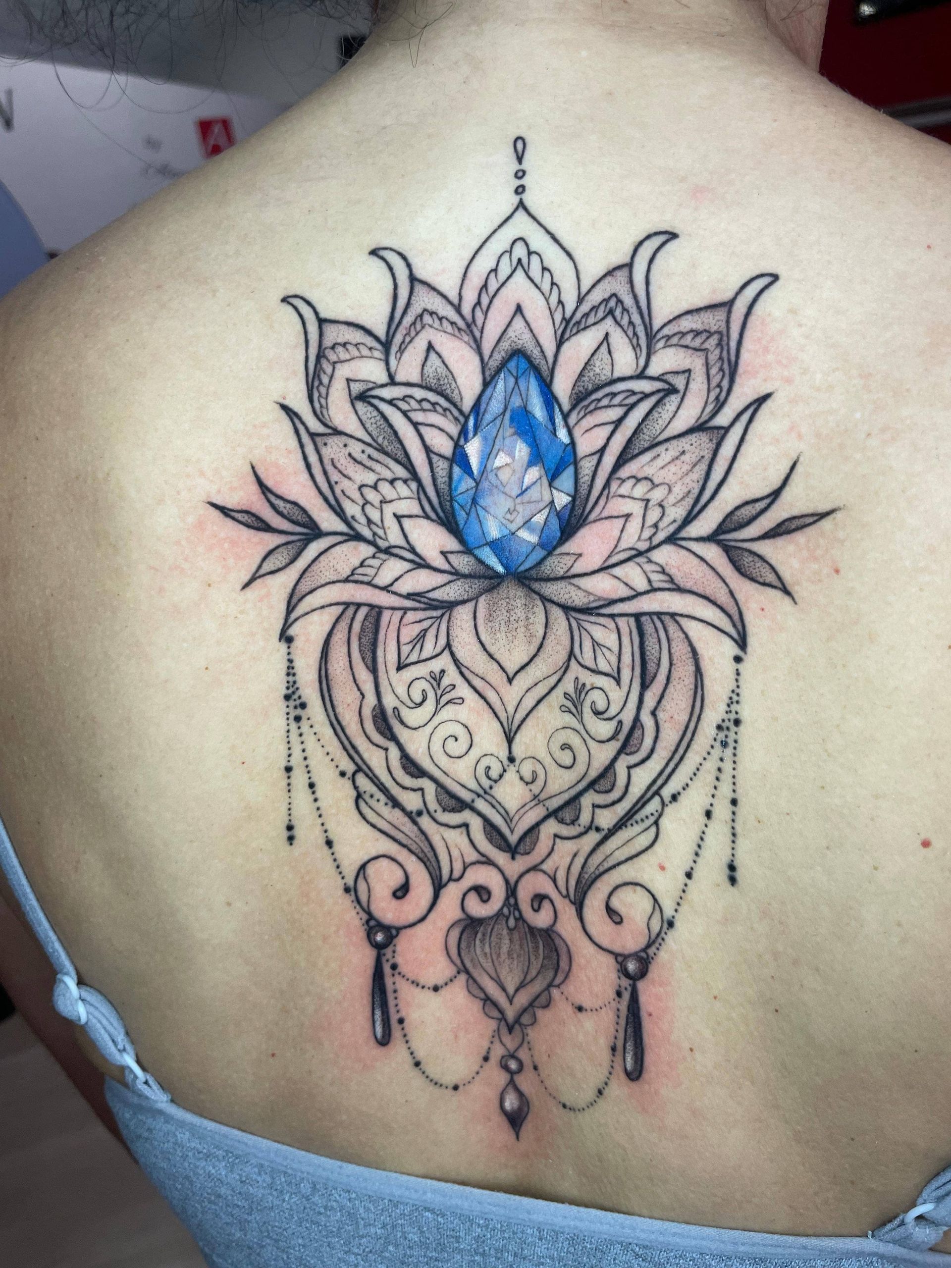 Tatuaje en la espalda de loto y mandala con una gema azul en forma de lágrima en el centro.
