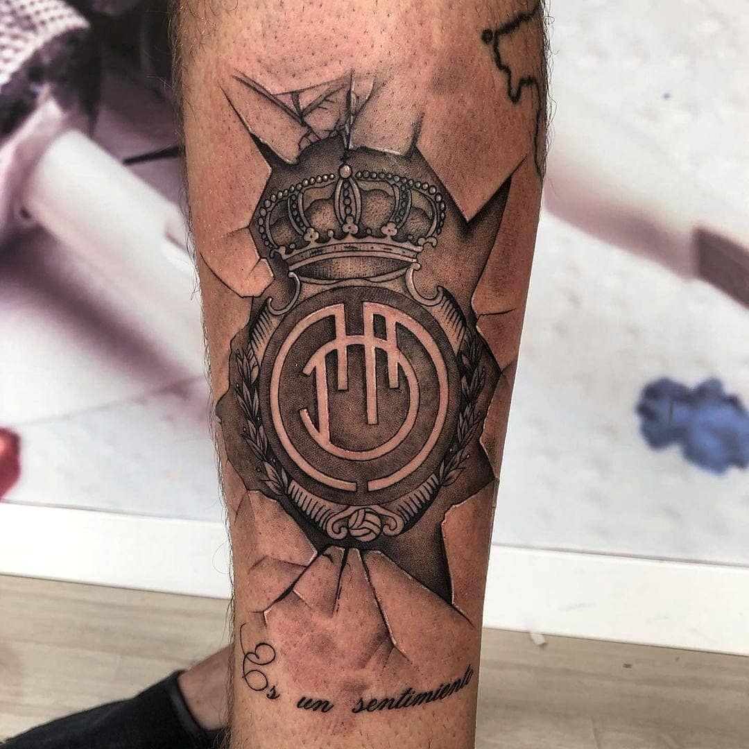 Tatuaje en blanco y negro en el antebrazo de un emblema circular coronado con líneas que parecen grietas y una pequeña inscripción debajo.