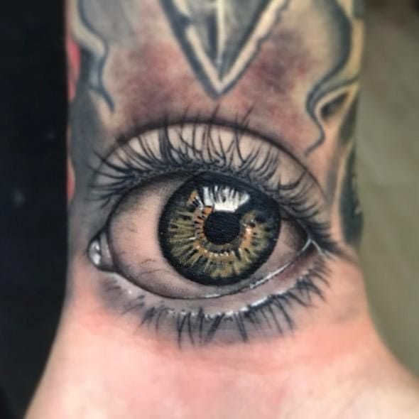 Primer plano de un tatuaje de ojo en la piel con iris verde-marrón y pestañas detalladas.