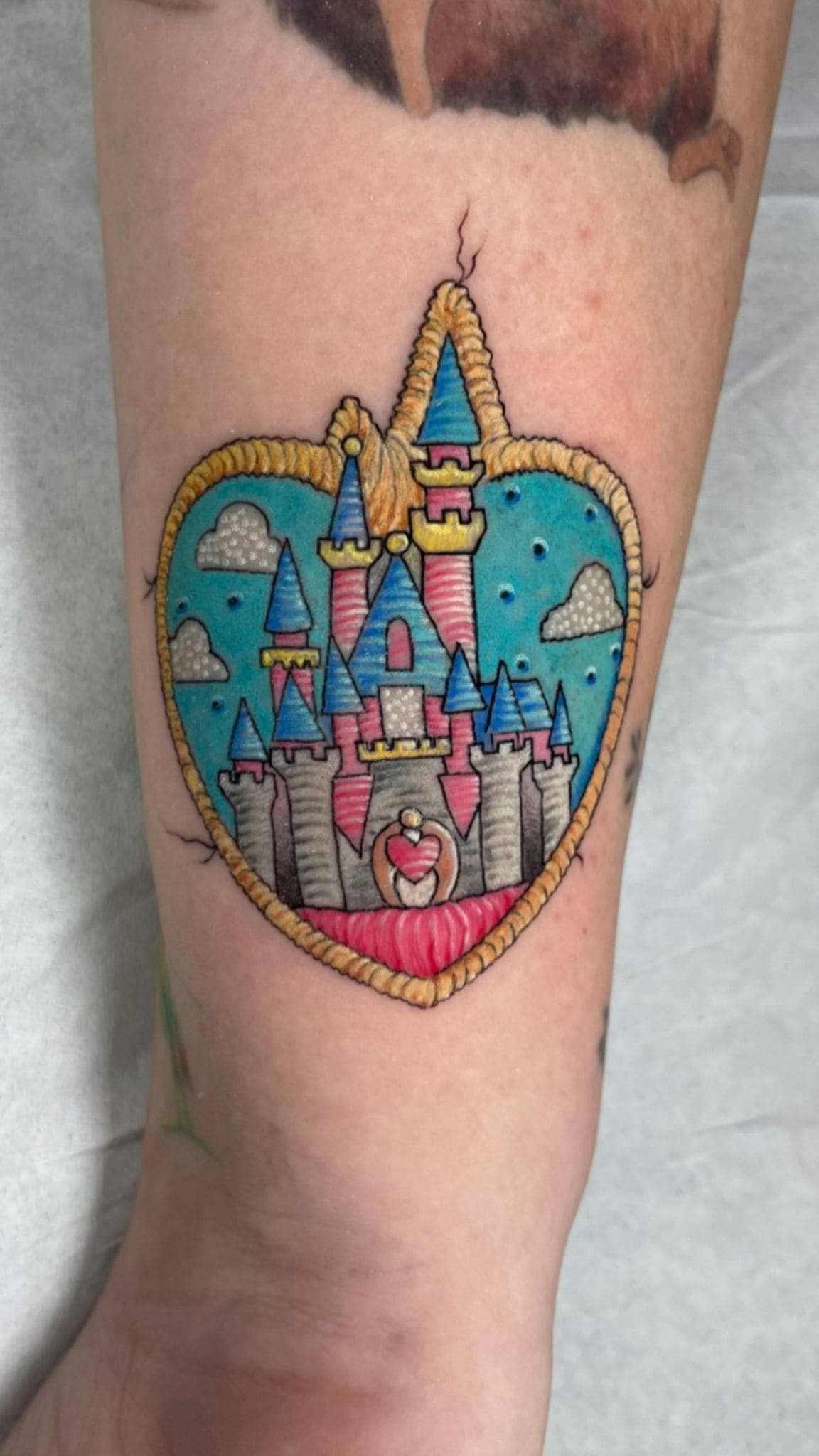 Tatuaje de un castillo colorido dentro de un corazón en la pantorrilla de una persona, con detalles en azul, rosa y amarillo.