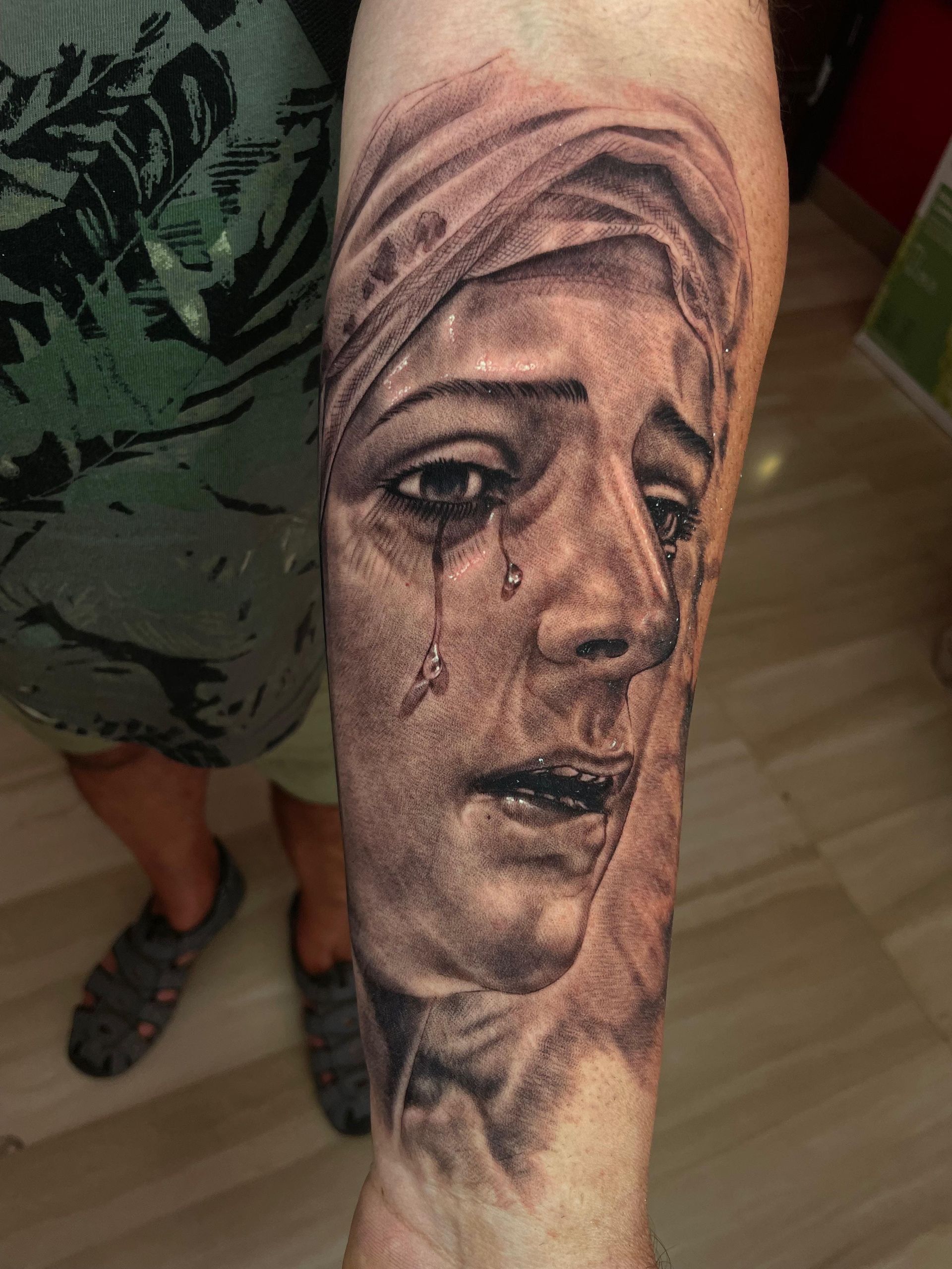 Primer plano de un tatuaje realista en blanco y negro de un rostro velado con una lágrima en un antebrazo.