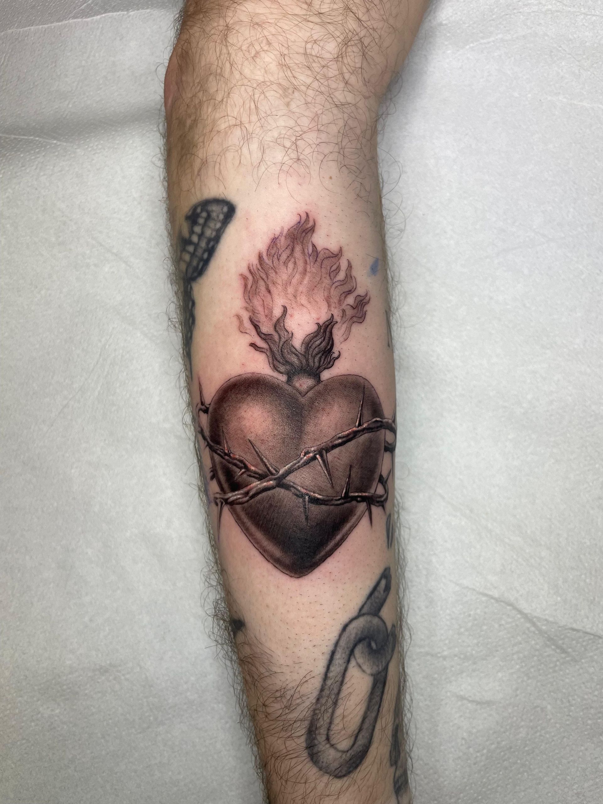 Un antebrazo tatuado con un corazón negro, una llama y diseños de líneas en bucle sobre una superficie clara.