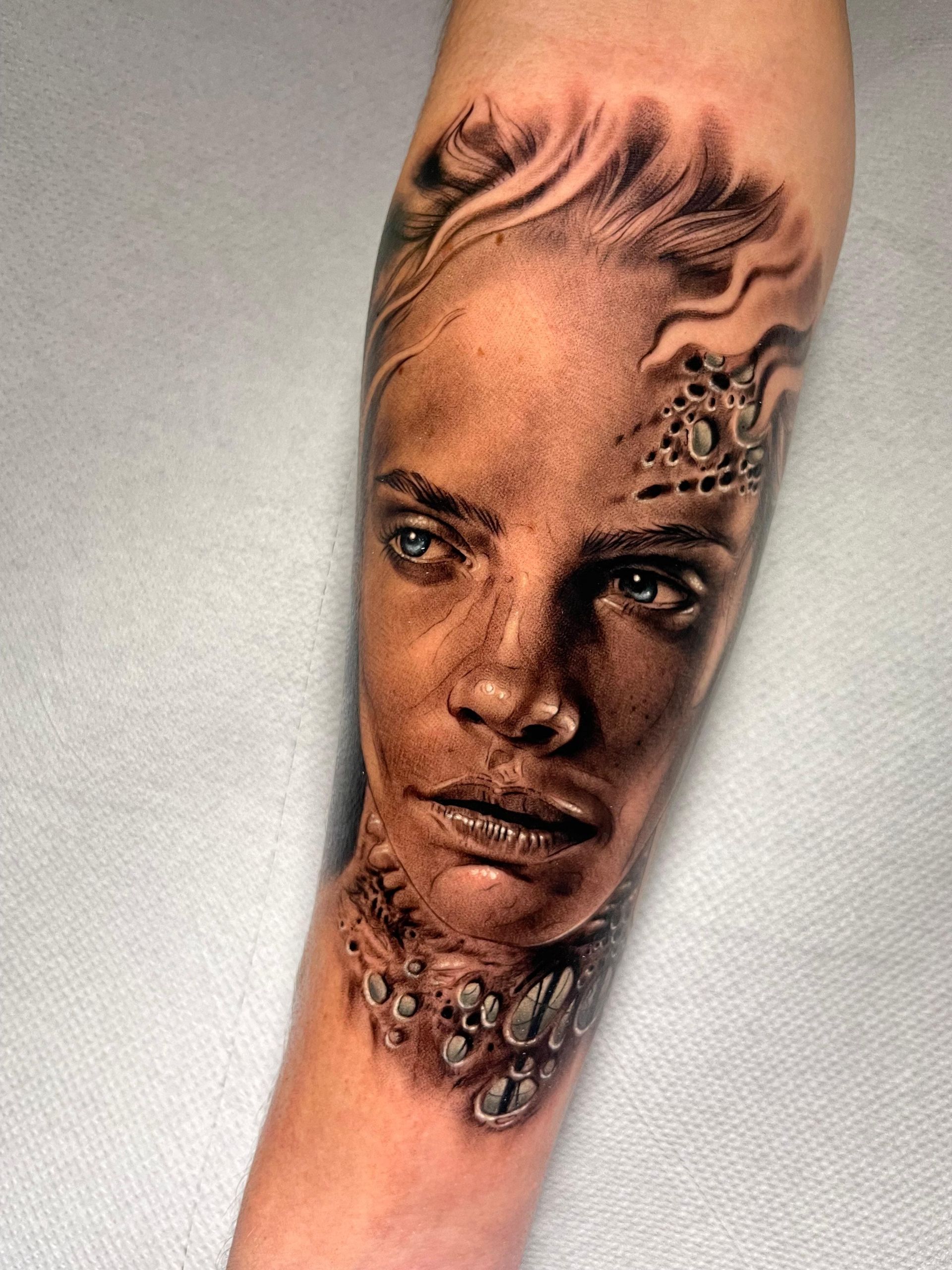 Tatuaje en primer plano de un rostro realista sobre la piel, con sombreado oscuro y detalles florales decorativos.