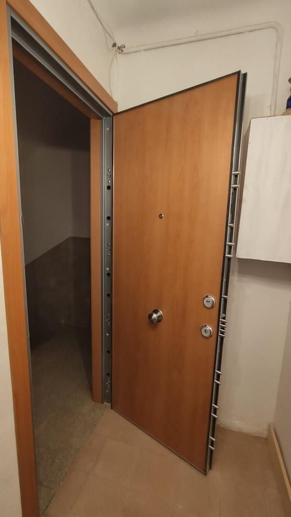 Puerta de seguridad marrón entreabierta en un pasillo, que revela una habitación clara. Herrajes cromados.