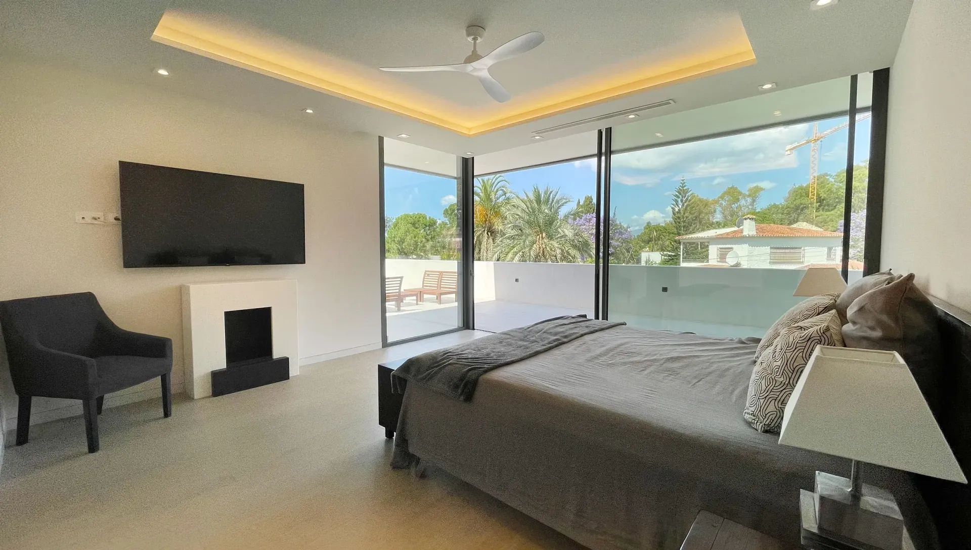 Dormitorio con amplios ventanales, cama, sillón, TV, chimenea y decoración moderna.