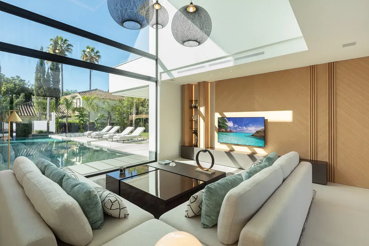 Moderna sala de estar con vista a la piscina, sofá blanco, ventanal y luces decorativas.