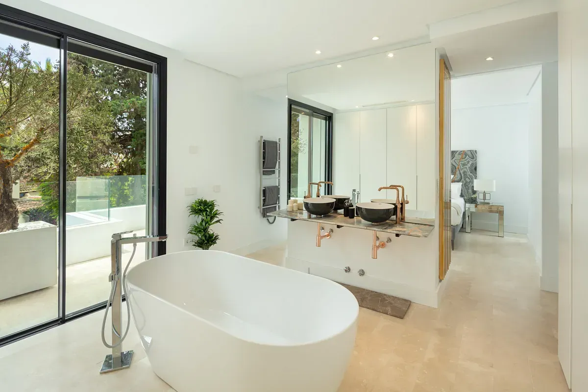 Baño moderno con bañera blanca, espejo grande y vista a la vegetación exterior.