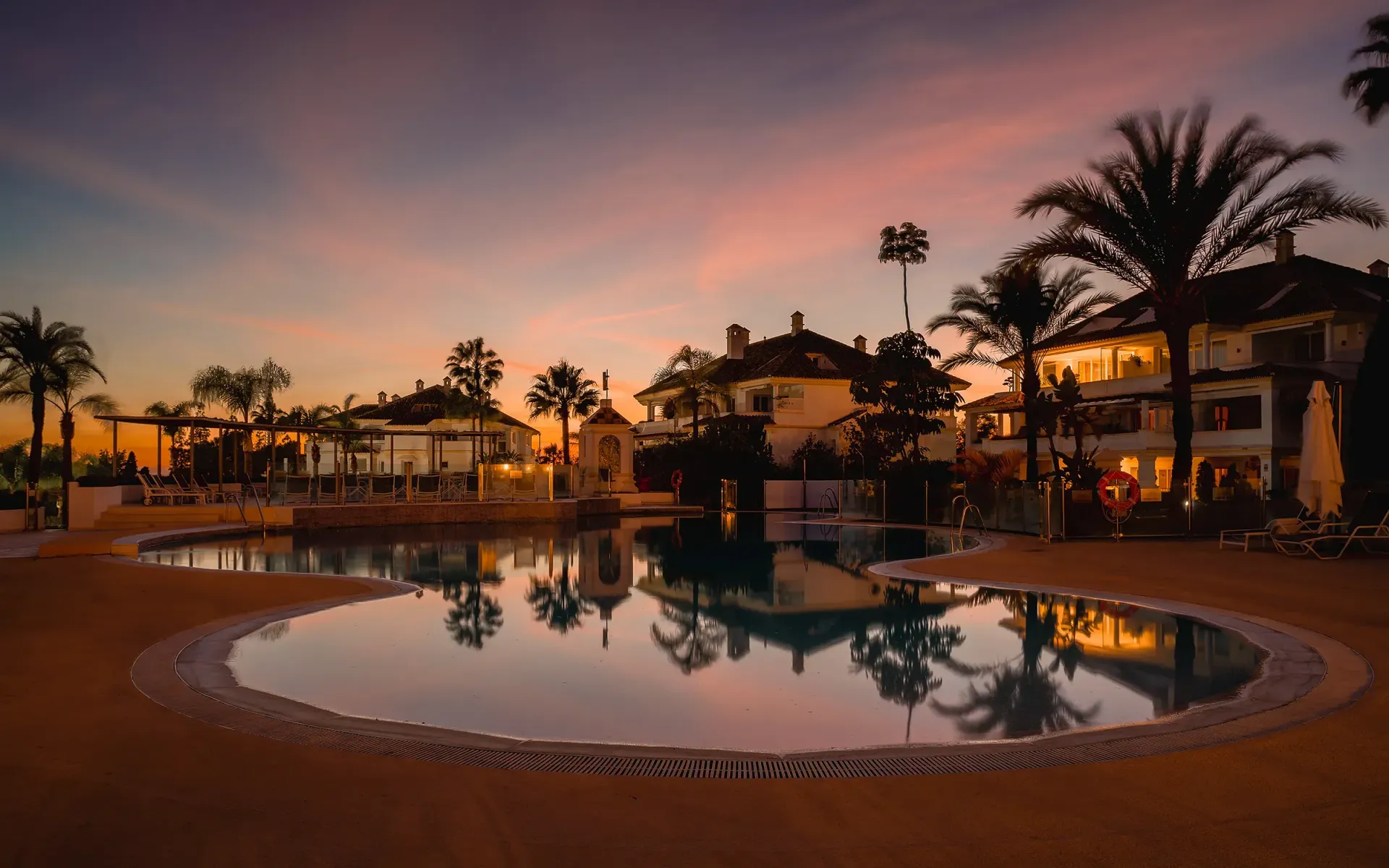 Piscina que refleja un colorido atardecer con palmeras y villas en un resort de lujo.