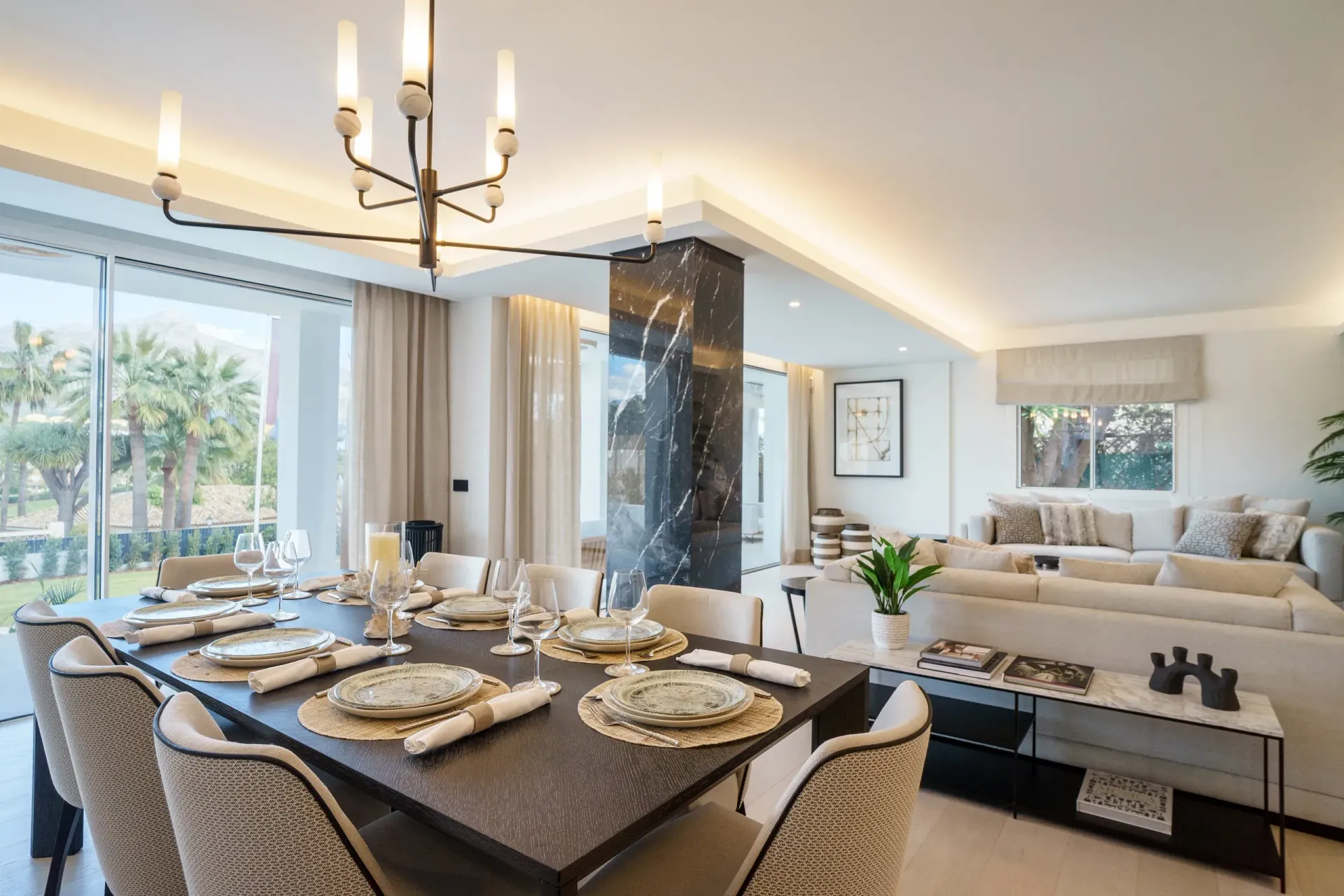 Elegante comedor con mesa negra preparada para comer, sala de estar adyacente con sofás neutros y vistas a las ventanas.