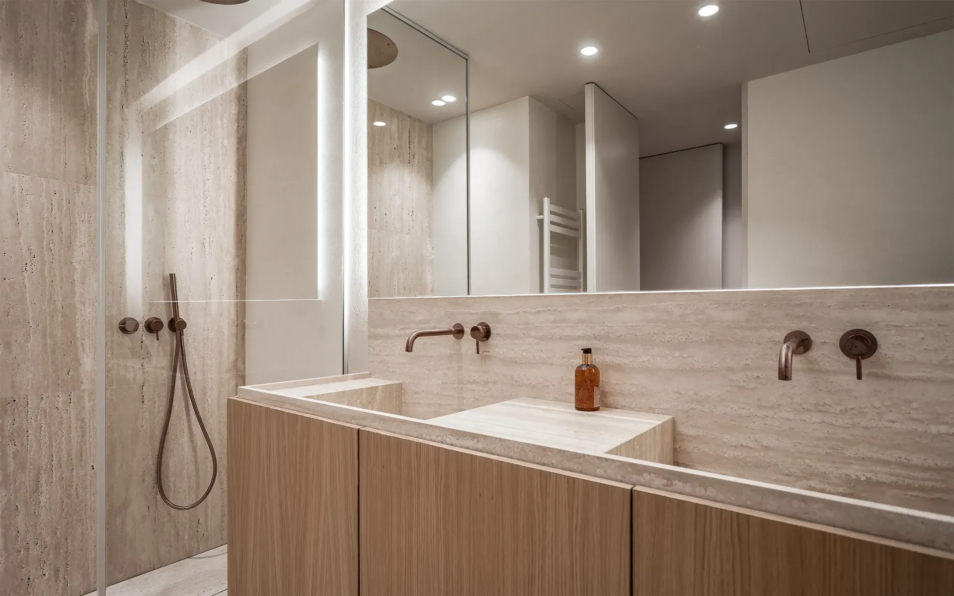 Baño moderno con paredes de piedra beige, muebles de madera clara y un espejo grande.
