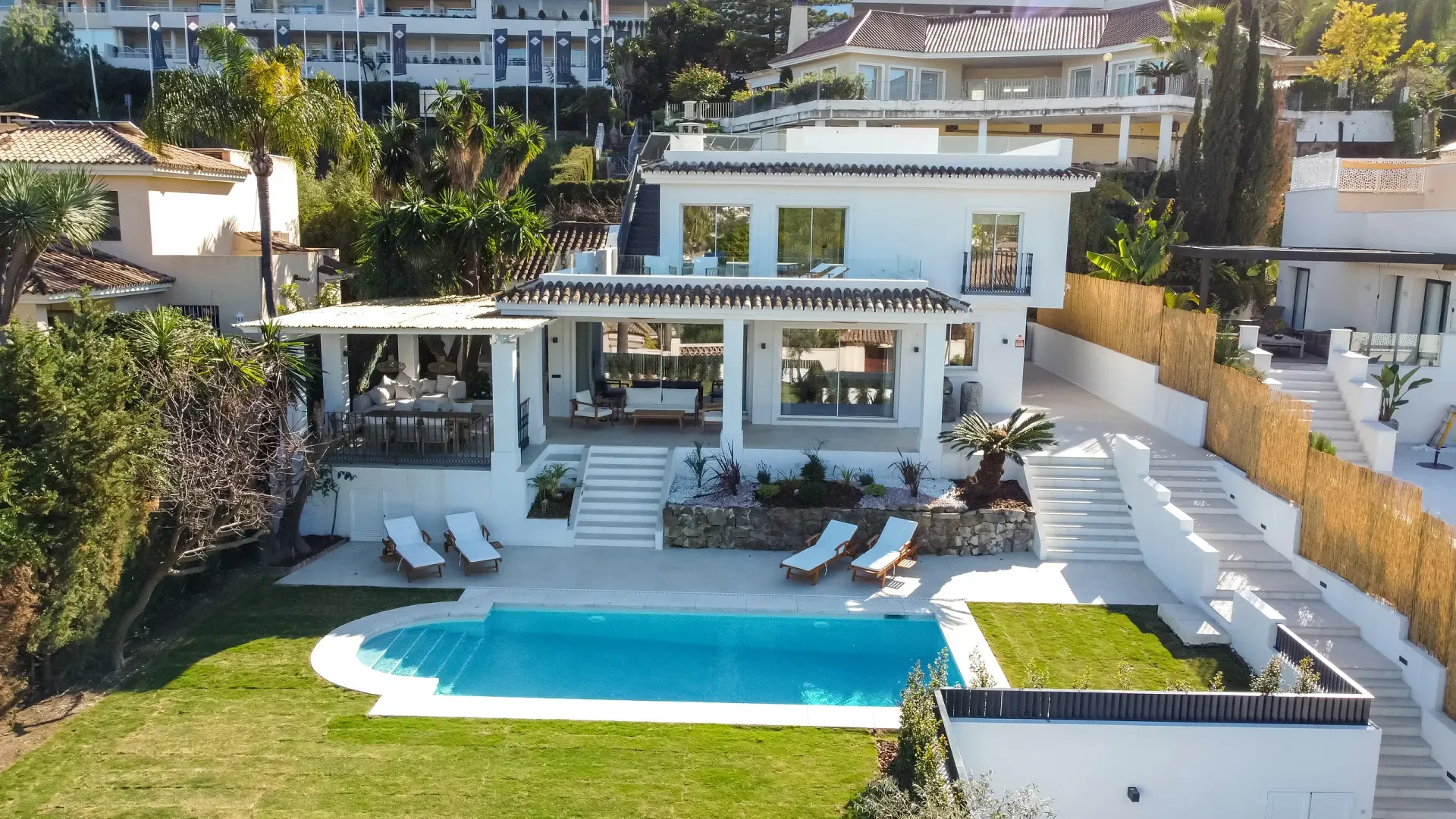 Villa blanca con piscina, césped exuberante y palmeras, situada en una zona soleada en la ladera.