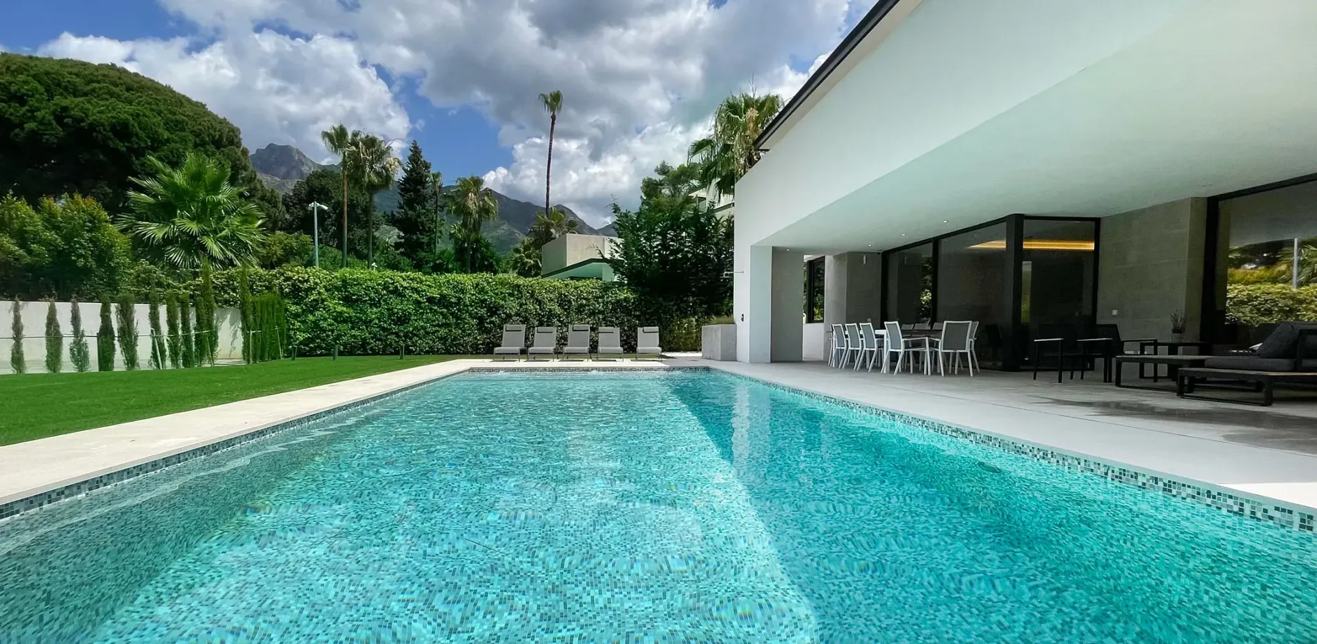 Una piscina junto a una moderna casa blanca con patio. Exuberante paisaje verde y cielo azul.