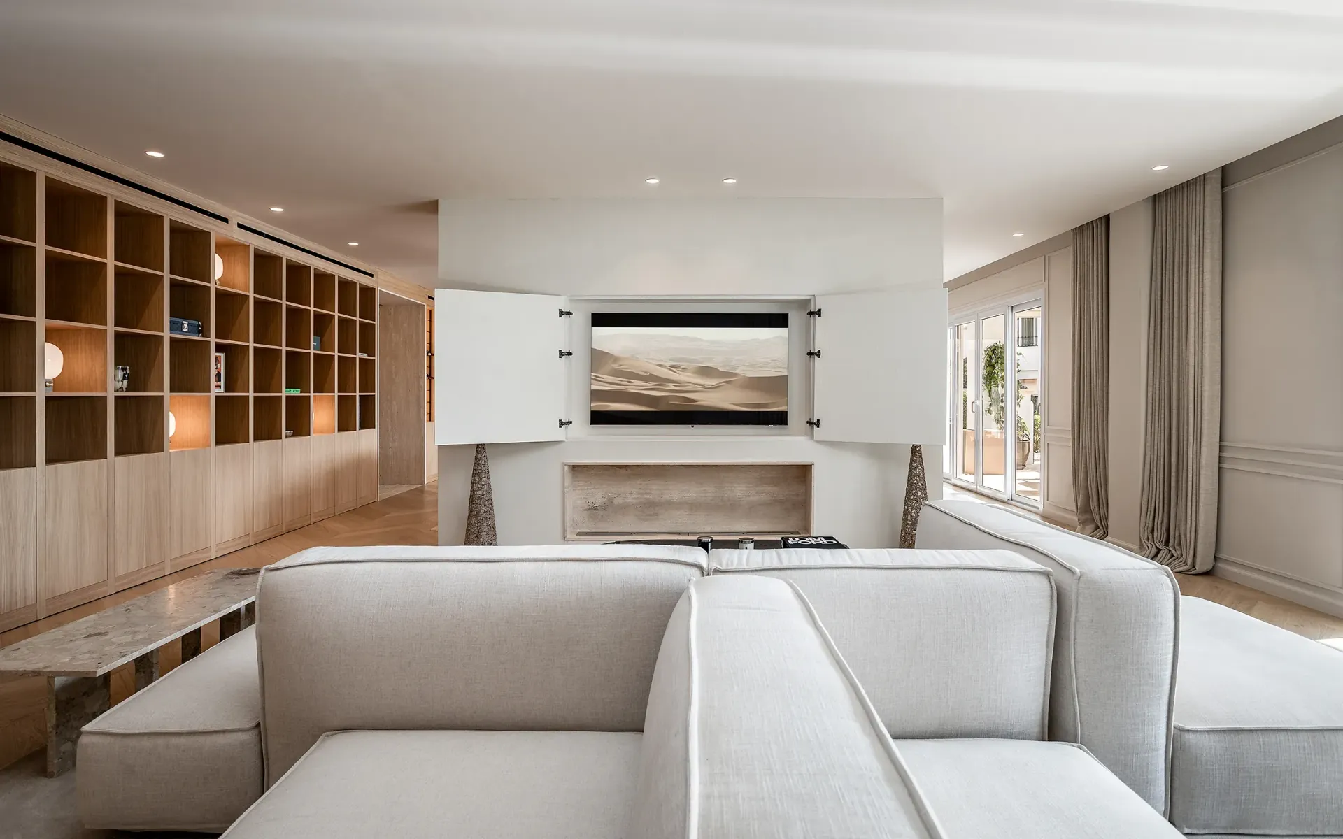 Sala de estar moderna en color beige con estantes integrados, un sofá seccional de color claro y un televisor oculto.