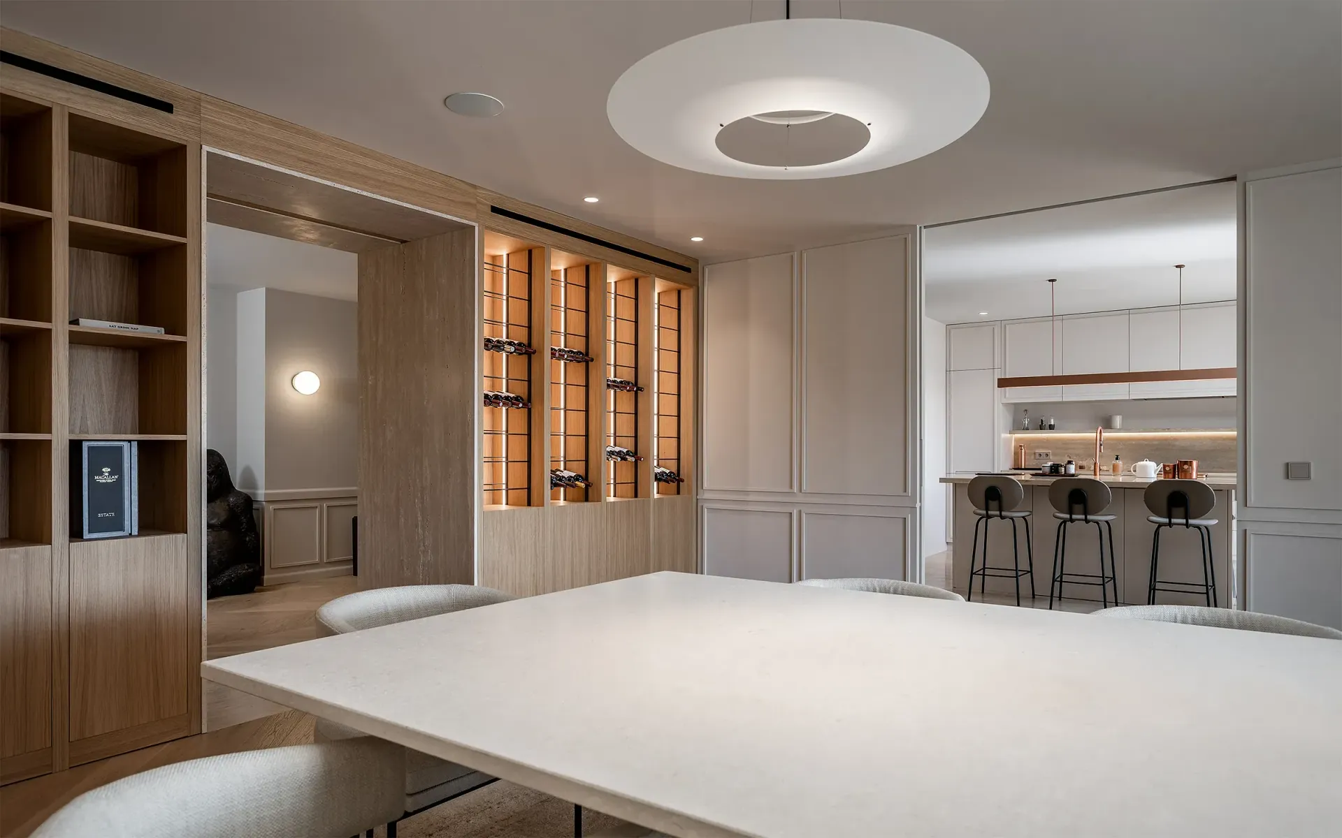 Comedor moderno con gran mesa blanca, estanterías abiertas, botellero y un vistazo a la cocina.