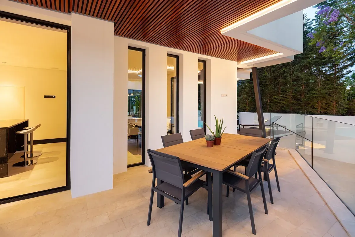 Comedor al aire libre con mesa de madera, sillas oscuras y un moderno techo blanco y marrón.
