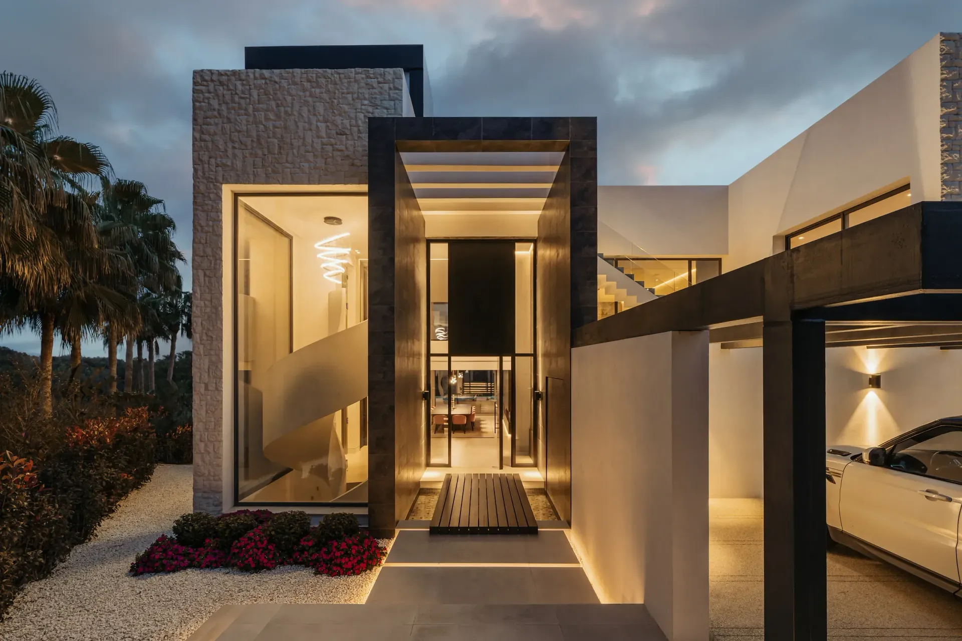 Exterior de villa moderna con entrada iluminada, paredes blancas y un coche en el garaje.