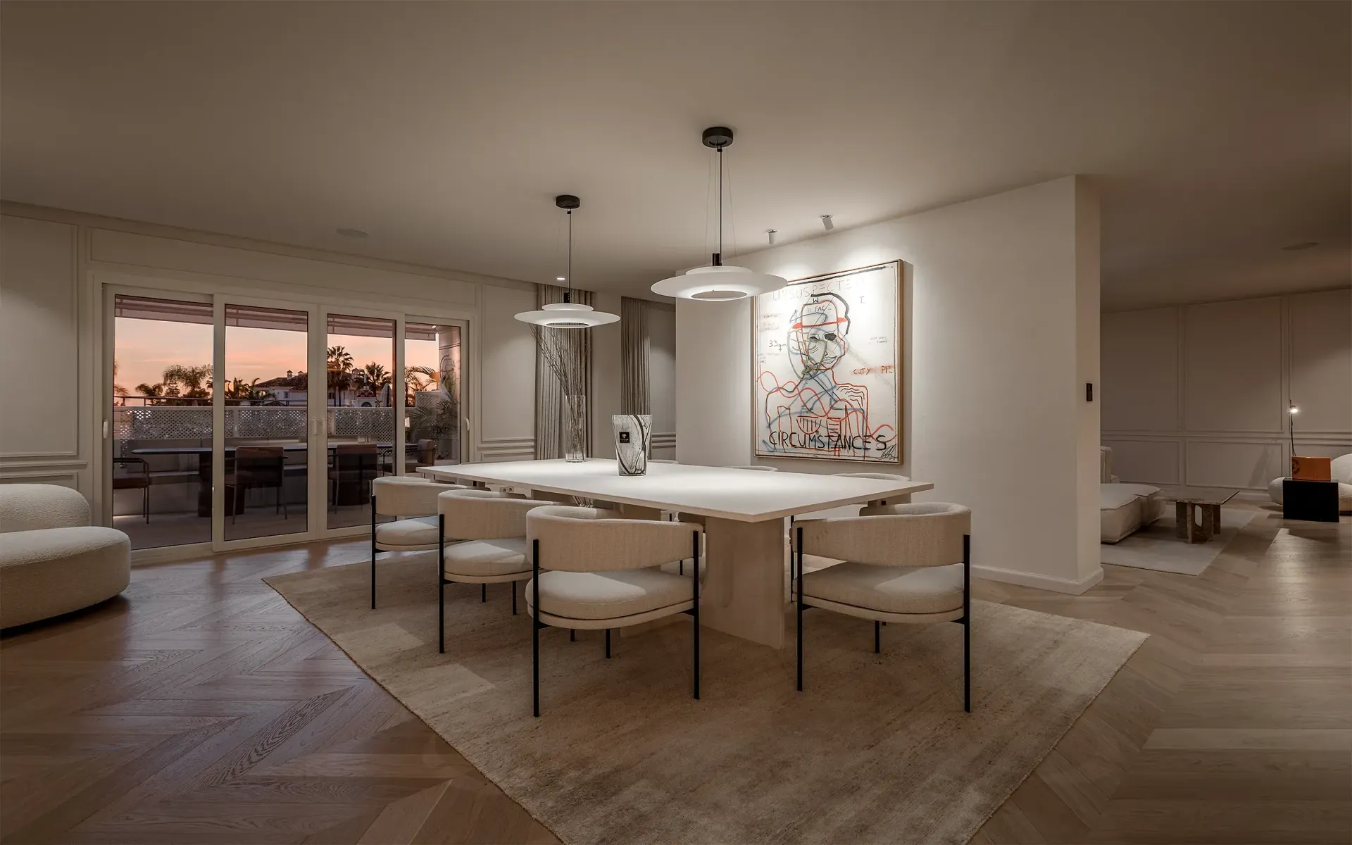 Comedor con mesa blanca, sillas y obras de arte. Puertas corredizas de vidrio ofrecen vistas del atardecer.