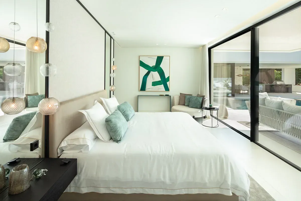 Amplio dormitorio blanco con cama grande, espejo de cuerpo entero, pared de vidrio con vista a los muebles del patio y a la piscina.