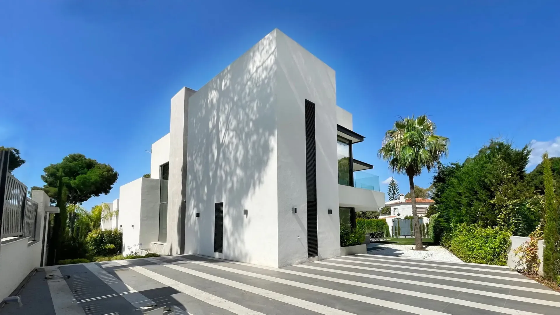 Casa blanca moderna con entrada gris y palmera contra un cielo azul.
