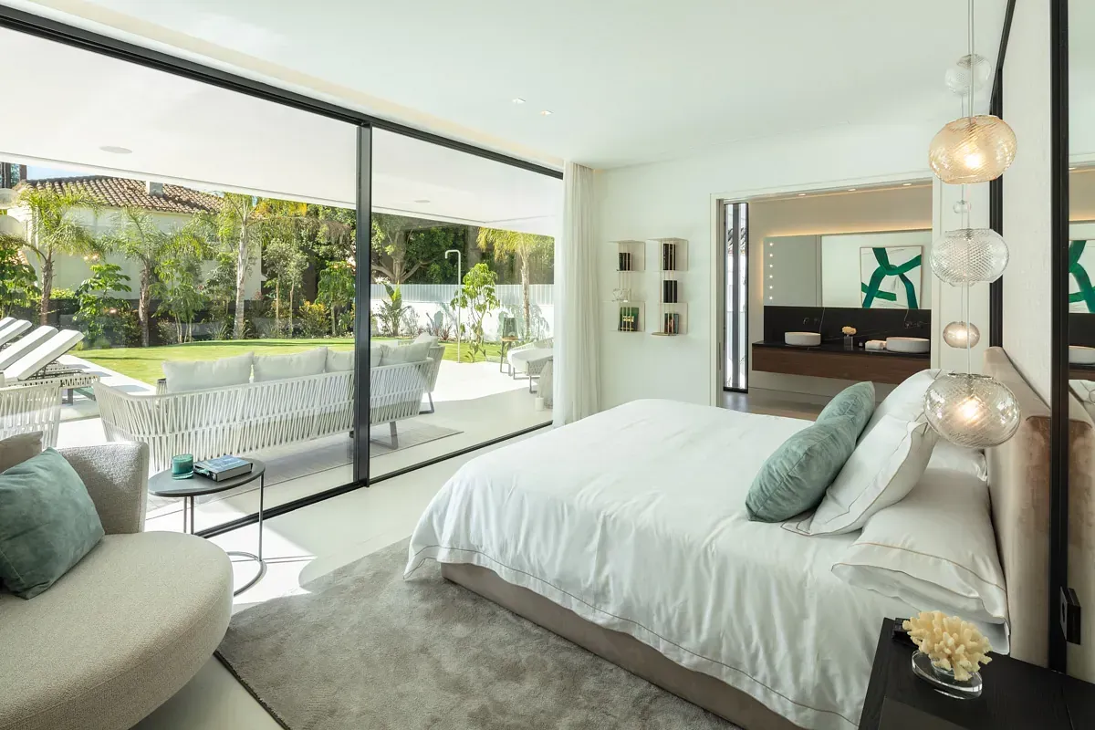 Dormitorio blanco y luminoso con puertas de cristal que dan a un patio y un jardín. Se ven la cama, los asientos y el baño.