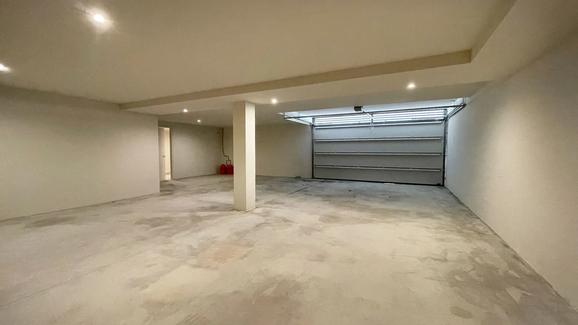 Habitación vacía, sin terminar, con piso de concreto, paredes blancas y puerta de garaje.