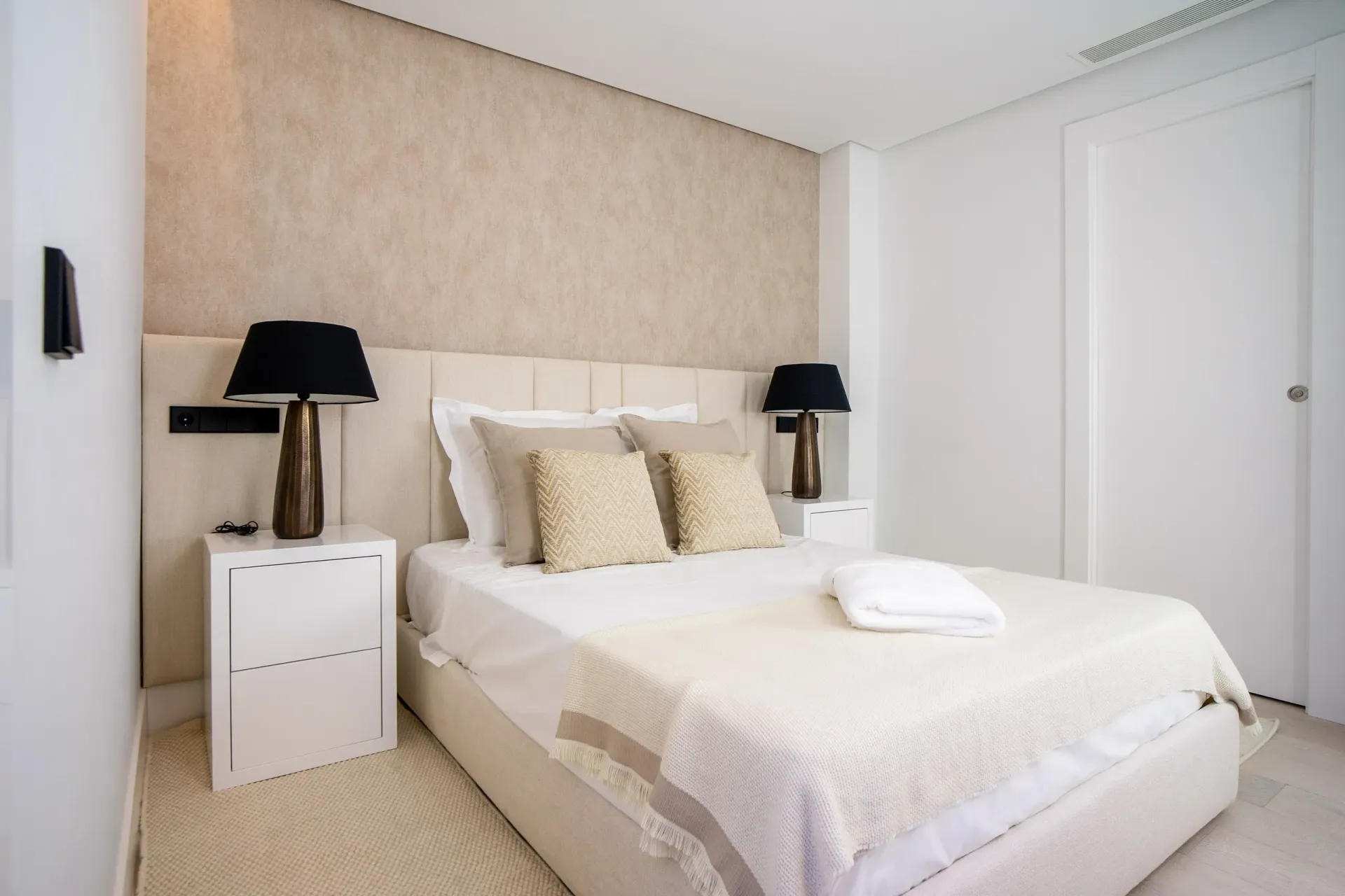 Dormitorio con pared texturizada beige, cama blanca, mesitas de noche, lámparas negras y puerta.