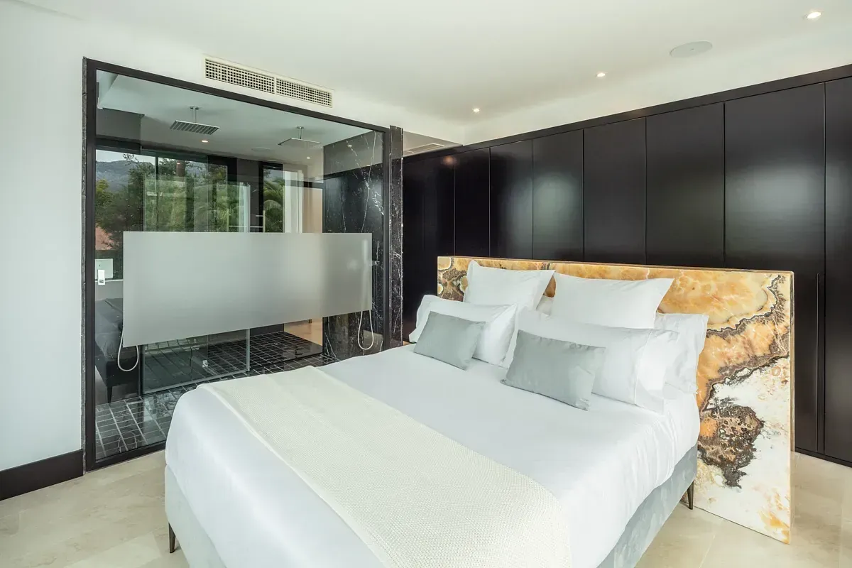 Dormitorio con una cama con cabecera ornamentada, ducha de vidrio esmerilado y paneles de madera oscura.