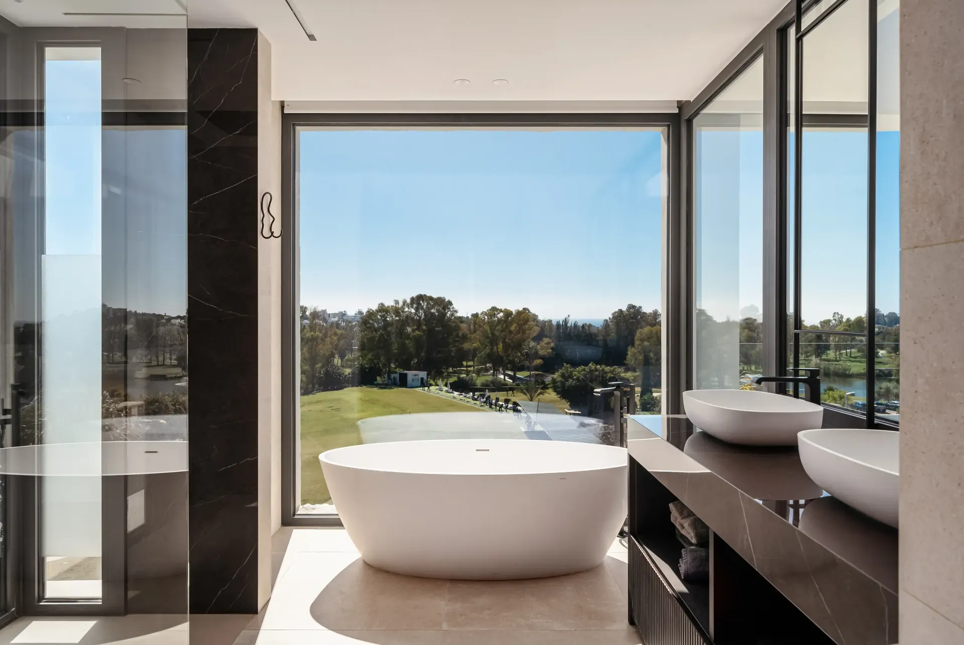 Baño moderno con un gran ventanal con vistas a un paisaje verde. Bañera blanca, lavabos y encimera oscura.