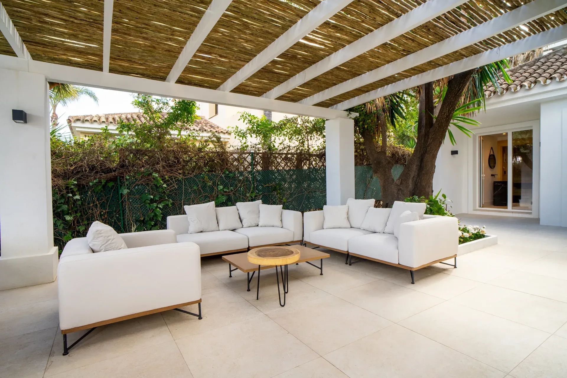 Zona de estar al aire libre con sofás blancos, techo tejido y follaje verde.