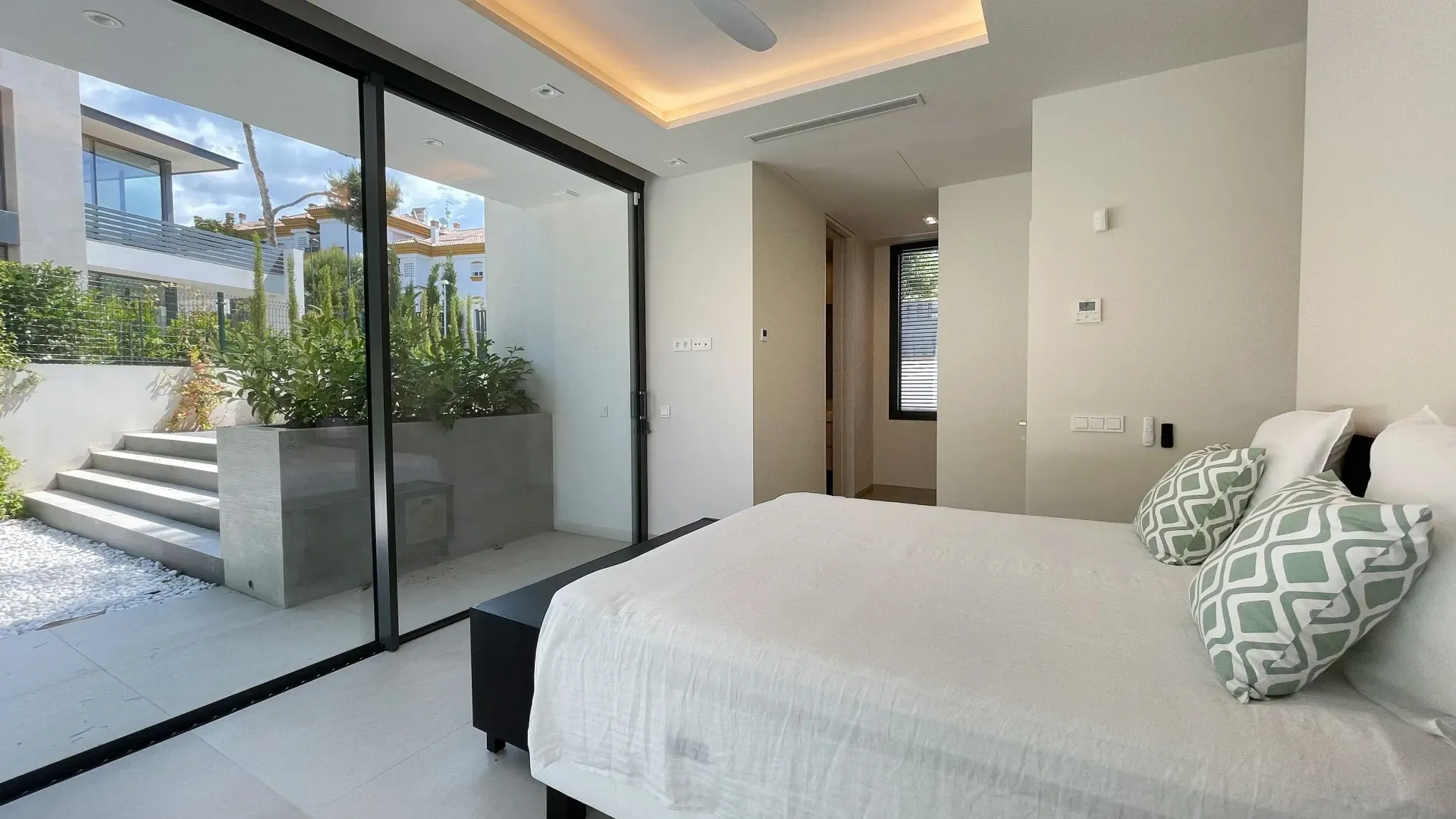 Dormitorio con gran puerta corrediza de vidrio, cama con almohadas verdes y vista a escaleras y paisaje.