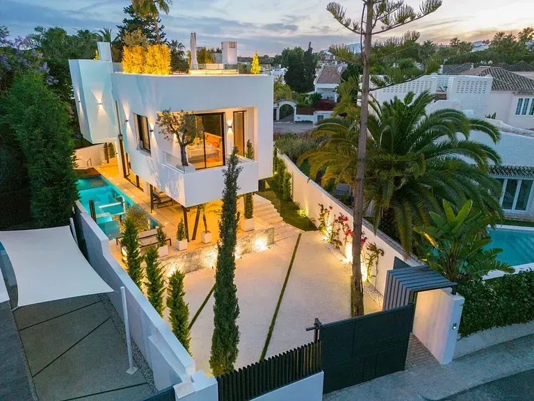 Moderna casa blanca con piscina, palmeras y entrada cerrada.