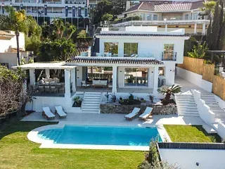 Villa blanca con piscina, tumbonas y escaleras, situada en césped verde.
