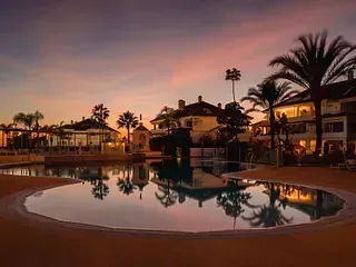 Una puesta de sol sobre la piscina de un resort, reflejando edificios y palmeras, creando una atmósfera tranquila.