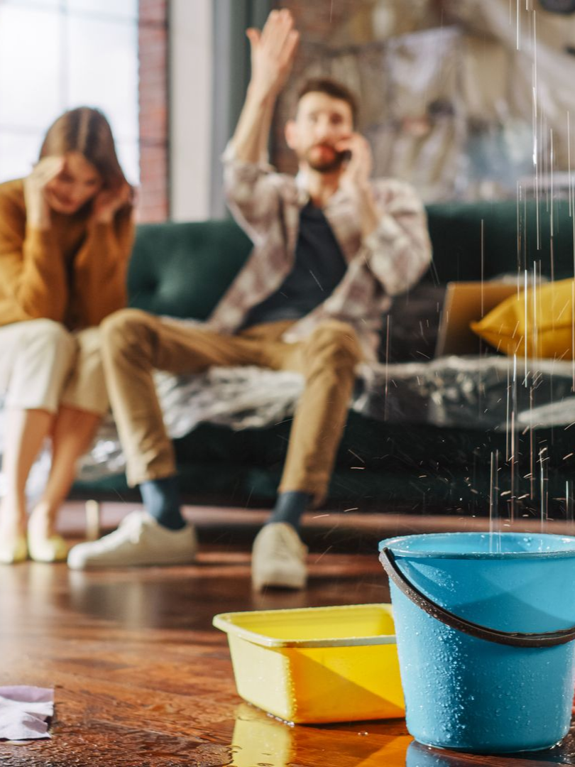 Intérieur d’un salon avec de l’eau qui tombe du plafond dans un seau, deux personnes assises sur un canapé 