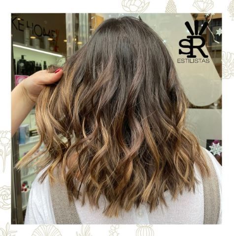 Cabello castaño ondulado con reflejos de una mujer. Una mano lo sujeta, posiblemente en una peluquería.