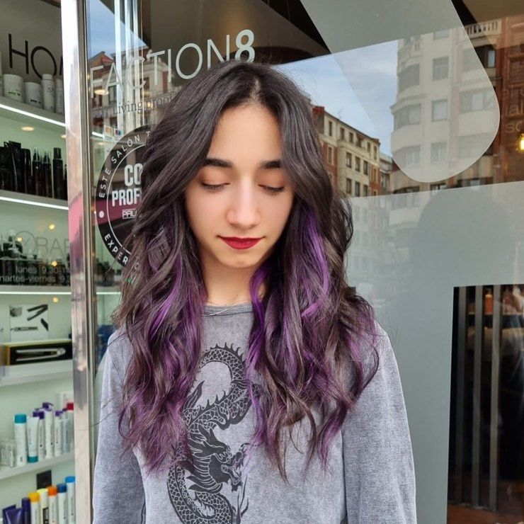 Mujer de cabello oscuro, reflejos morados y sudadera gris. De pie frente a una peluquería.