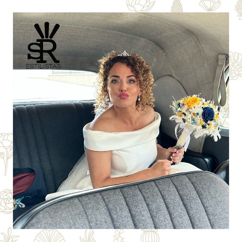 Novia con vestido blanco frunciendo los labios y sosteniendo un ramo en un coche clásico.