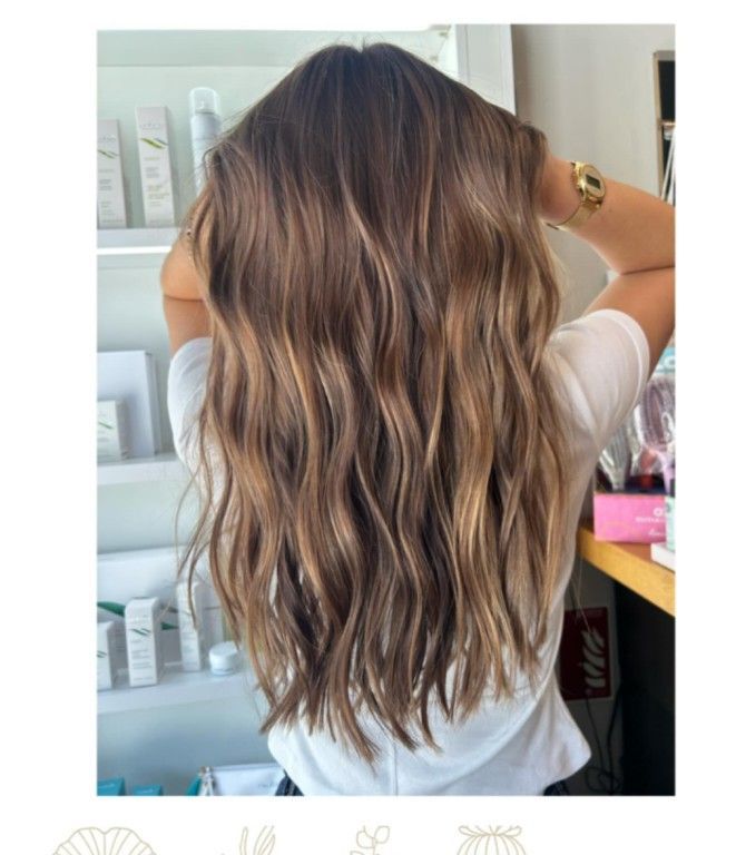 Mujer con cabello castaño largo y ondulado con reflejos rubios, de pie frente a estantes con productos.