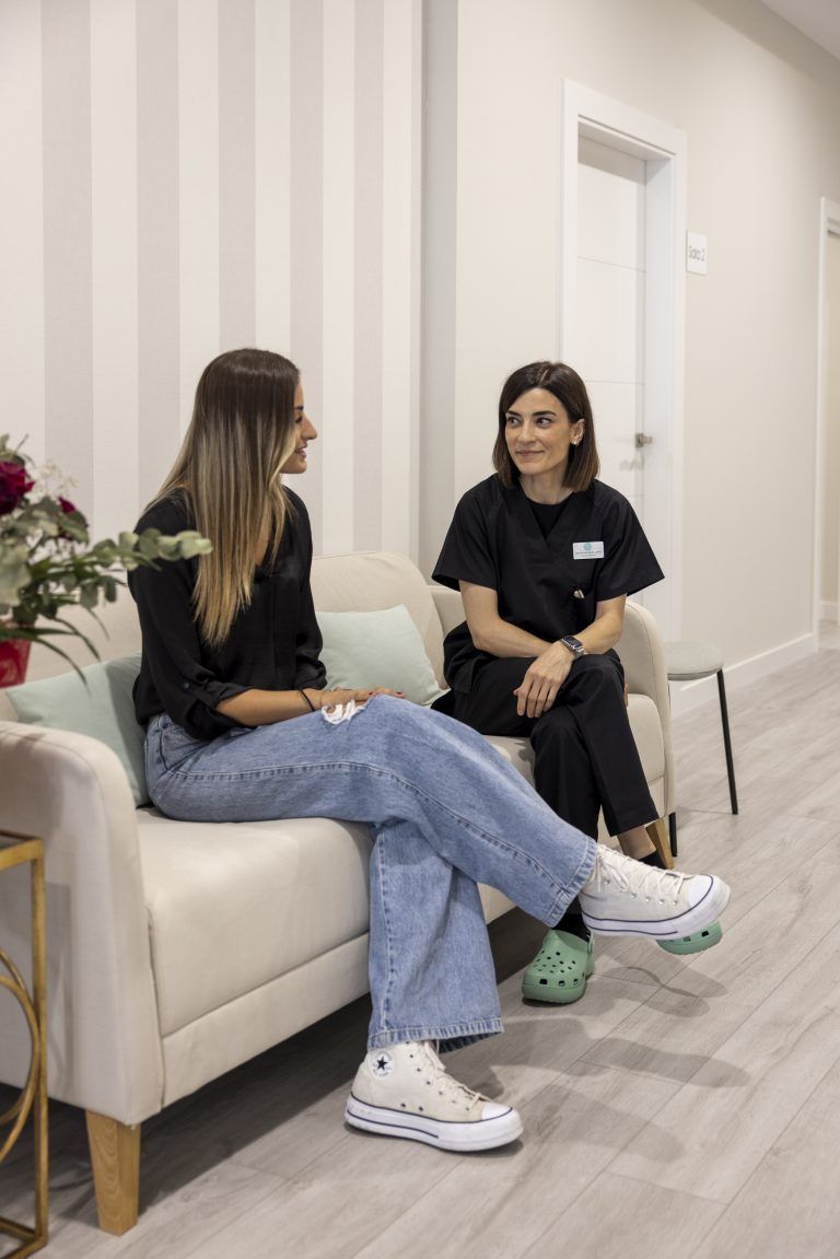 Dos mujeres están sentadas en un sofá en una sala de espera hablando entre sí.