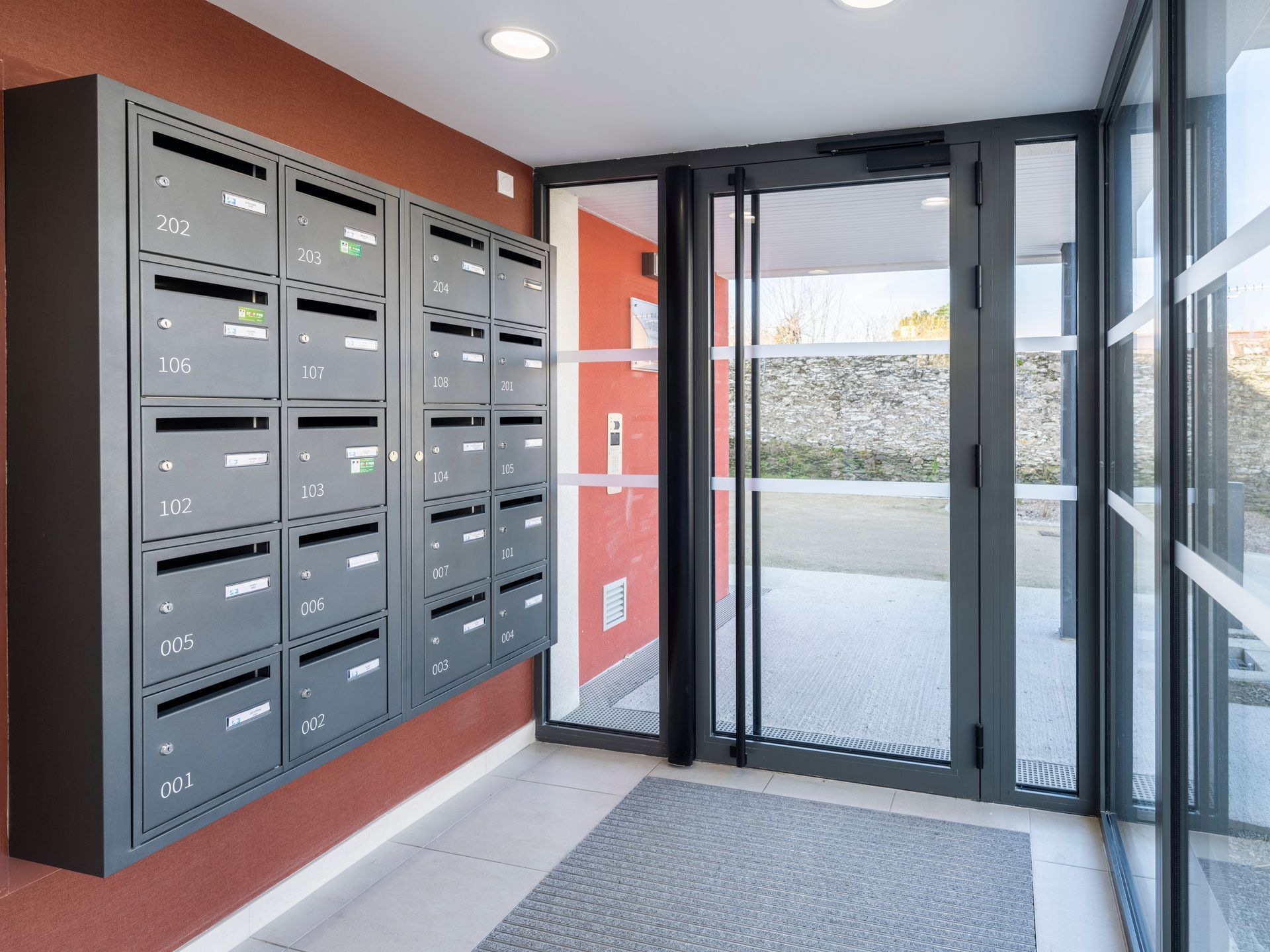 Boîtes aux lettres dans le hall d'un immeuble, à côté d'une porte d'entrée vitrée.